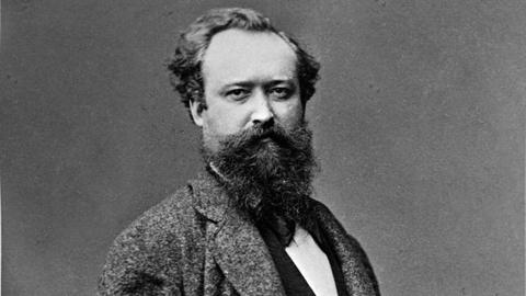 Schwarzweißporträt von Wilhelm Busch, der an eine Wand gelehnt in die Kamera blickt.