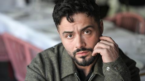 Der Schauspieler Aram Arami schaut offen in die Kamera und hält eine Hand an seinem Gesicht.