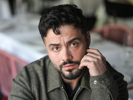 Der Schauspieler Aram Arami schaut offen in die Kamera und hält eine Hand an seinem Gesicht.