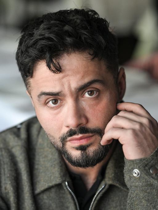Der Schauspieler Aram Arami schaut offen in die Kamera und hält eine Hand an seinem Gesicht.