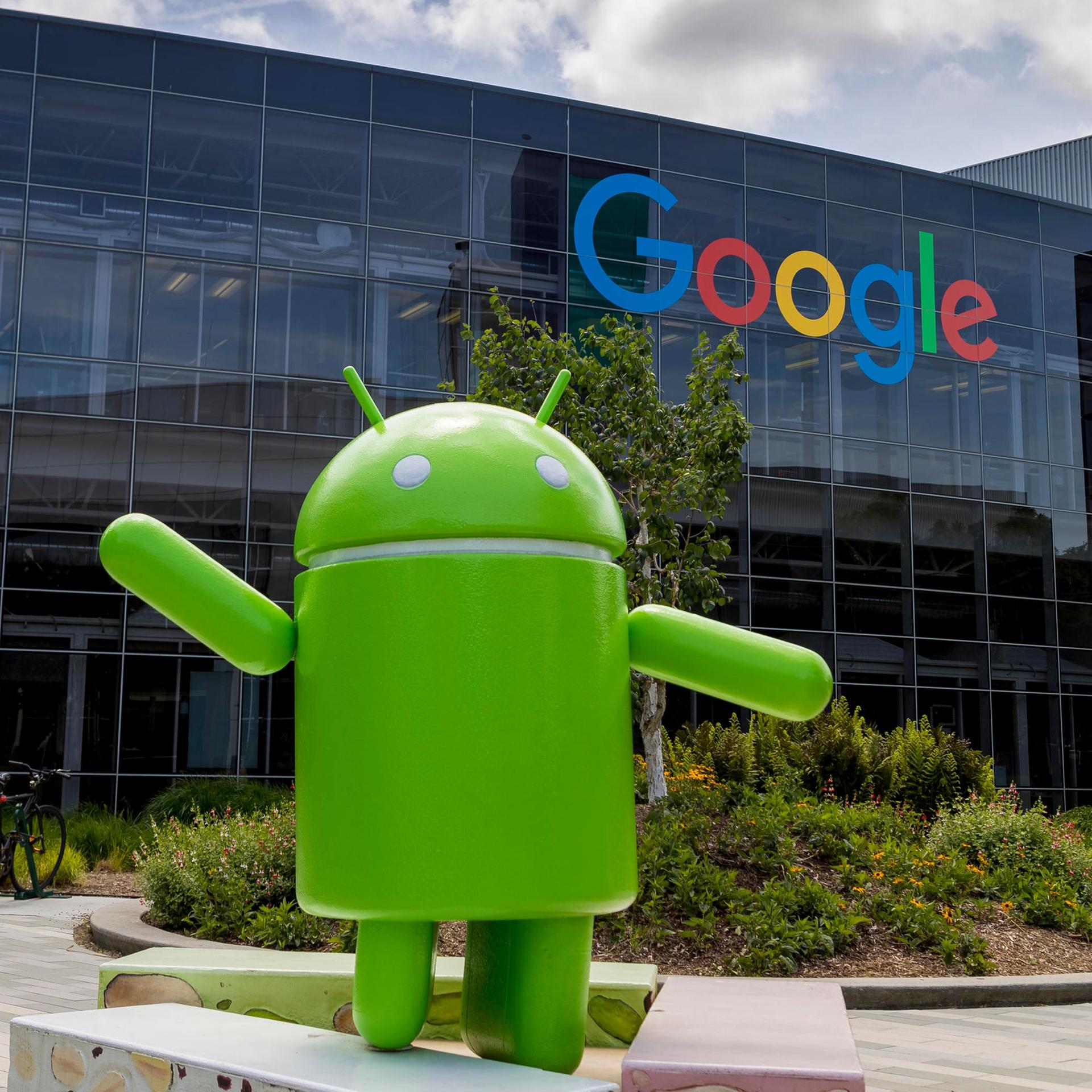 Blick auf die gläserne Fassade mit Google Logo beim Firmensitz in Mountain View, Kalifornien. Davor steht ein grünes Tonnenmännchen.