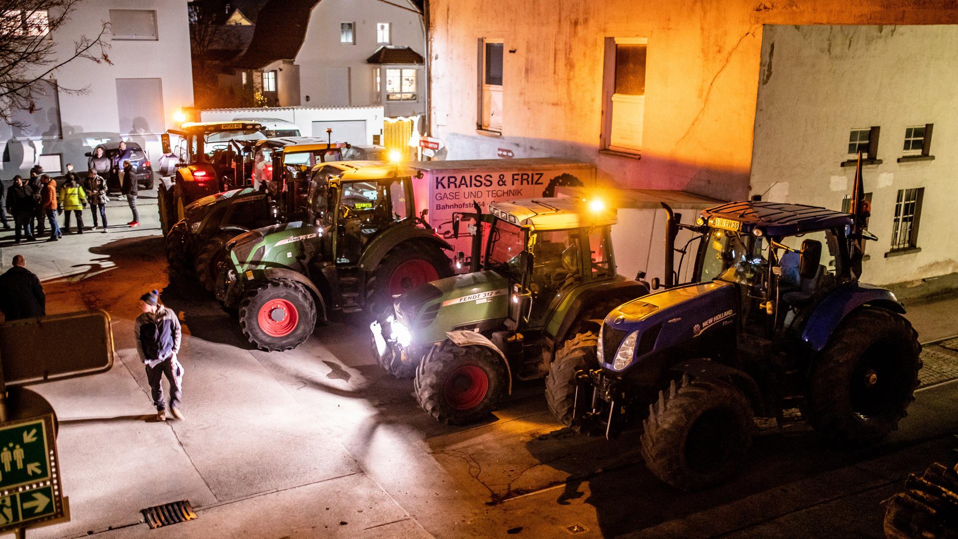 Es ist dunkel. Fünf Traktoren mit eingeschalteten Scheinwerfern stehen vor einem Haus. Drumherung mehrere Menschen.