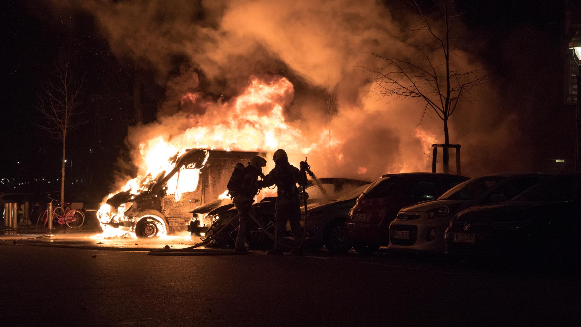 Feuerwehrleute versuchen ein brennendes Auto zu löschen. Es ist Nacht und alles ist sehr dunkel, nur das Feuer sticht hervor.