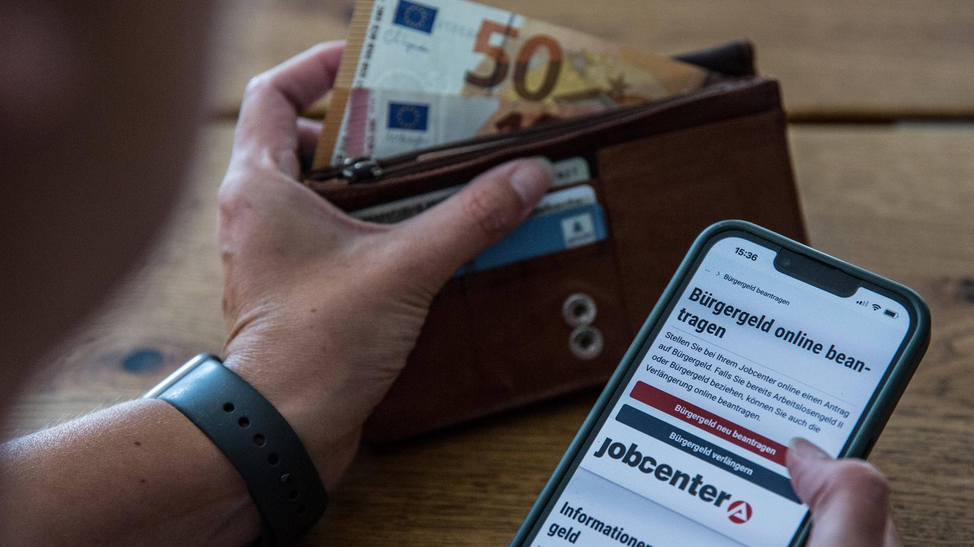 Ein Symbolfoto zeigt die Hand einer Person, die am Tisch sitzt und sich auf dem Smartphone über den Antrag für Bürgergeld informiert. In der anderen Hand hält sie eine Geldbörse, aus der Geldscheine ragen.