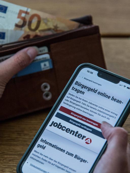 Ein Symbolfoto zeigt die Hand einer Person, die am Tisch sitzt und sich auf dem Smartphone über den Antrag für Bürgergeld informiert. In der anderen Hand hält sie eine Geldbörse, aus der Geldscheine ragen.