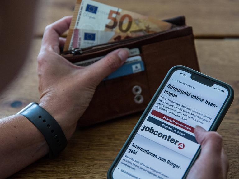 Ein Symbolfoto zeigt die Hand einer Person, die am Tisch sitzt und sich auf dem Smartphone über den Antrag für Bürgergeld informiert. In der anderen Hand hält sie eine Geldbörse, aus der Geldscheine ragen.
