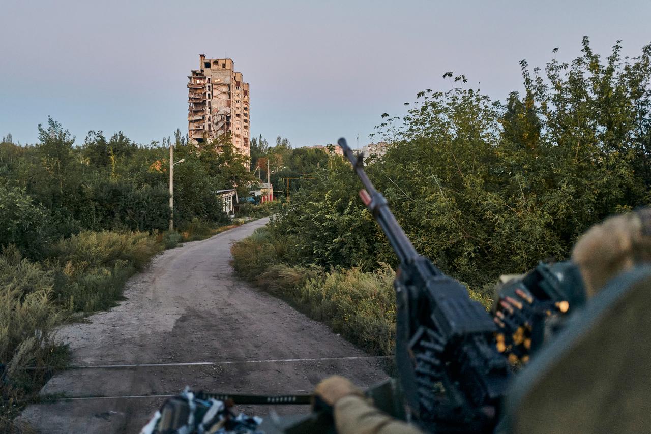 Ein Maschinengewehr ragt im Vordergrund unscharf ins Bild, im Hintergrund ist ein einzelnes Hochhaus vor einem Feldweg zu sehen.