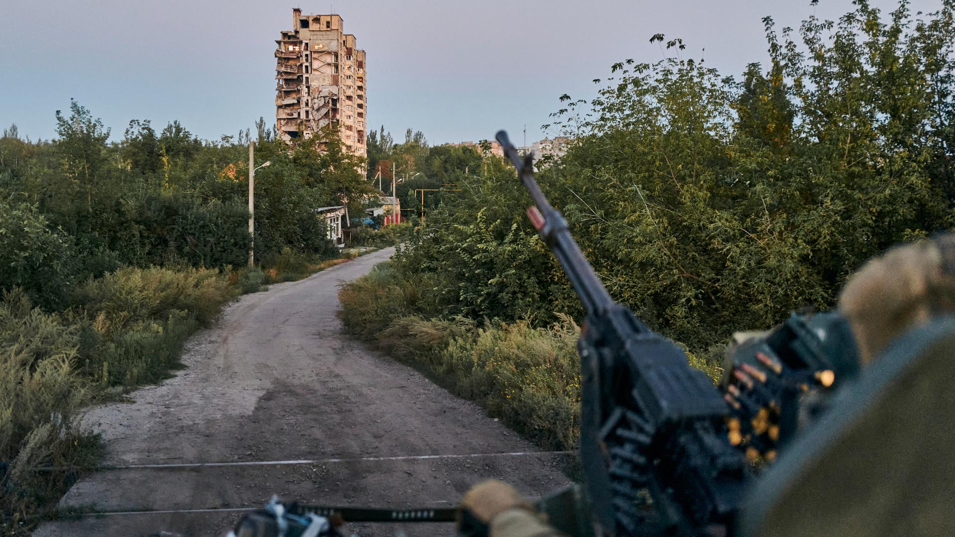 Ein Maschinengewehr ragt im Vordergrund unscharf ins Bild, im Hintergrund ist ein einzelnes Hochhaus vor einem Feldweg zu sehen.