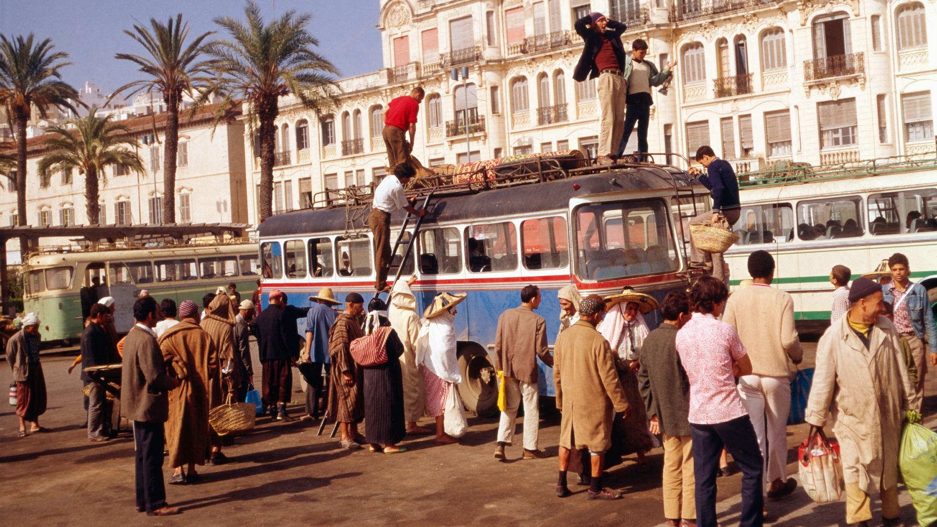 Das Foto zeigt die Stadt Tanger in den 1980er Jahren. Mehrere Menschen stehen um einen Reisebus, ein paar verladen Gepäck auf das Dach.