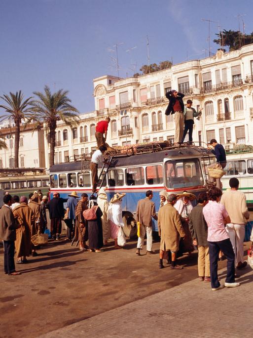 Das Foto zeigt die Stadt Tanger in den 1980er Jahren. Mehrere Menschen stehen um einen Reisebus, ein paar verladen Gepäck auf das Dach.