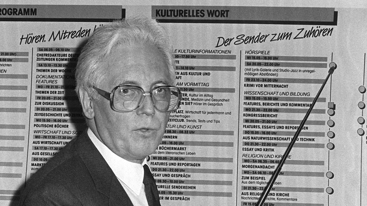 Richard Becker steht in einem Schwarz-Weiß-Bild vor einer Tafel mit der Aufschrift "Deutschlandfunk".