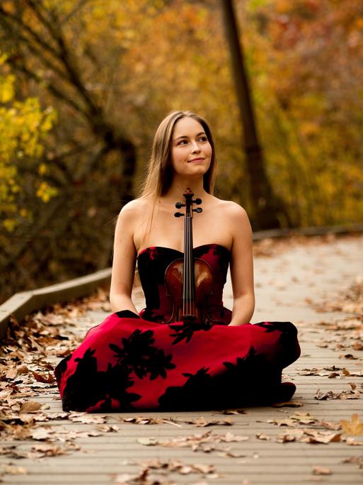 Geneva Lewis sitzt mit ihrer Geige im Schos in einem Abendkleid auf einem gepflasterten Weg, der von herbstlichen Bäumen umgeben ist.