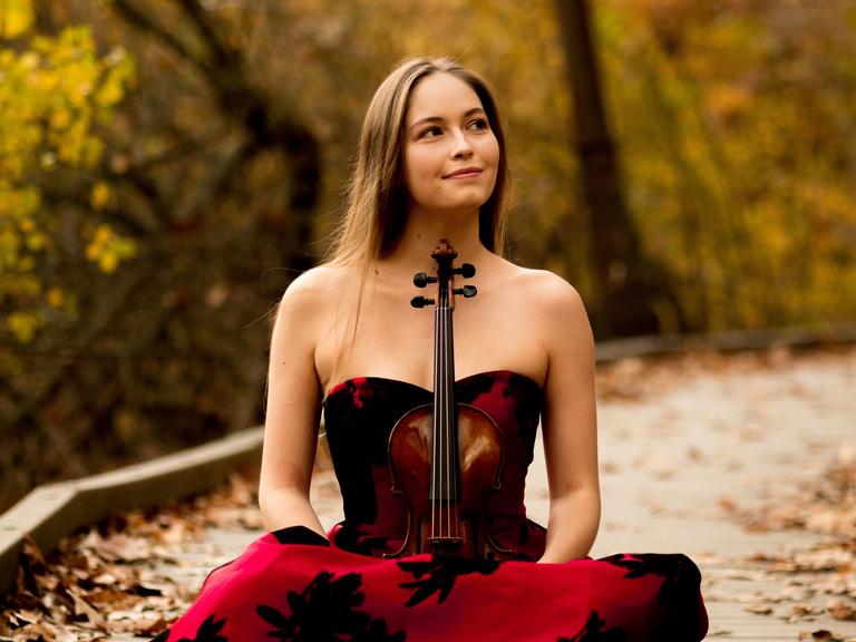 Geneva Lewis sitzt mit ihrer Geige im Schos in einem Abendkleid auf einem gepflasterten Weg, der von herbstlichen Bäumen umgeben ist.