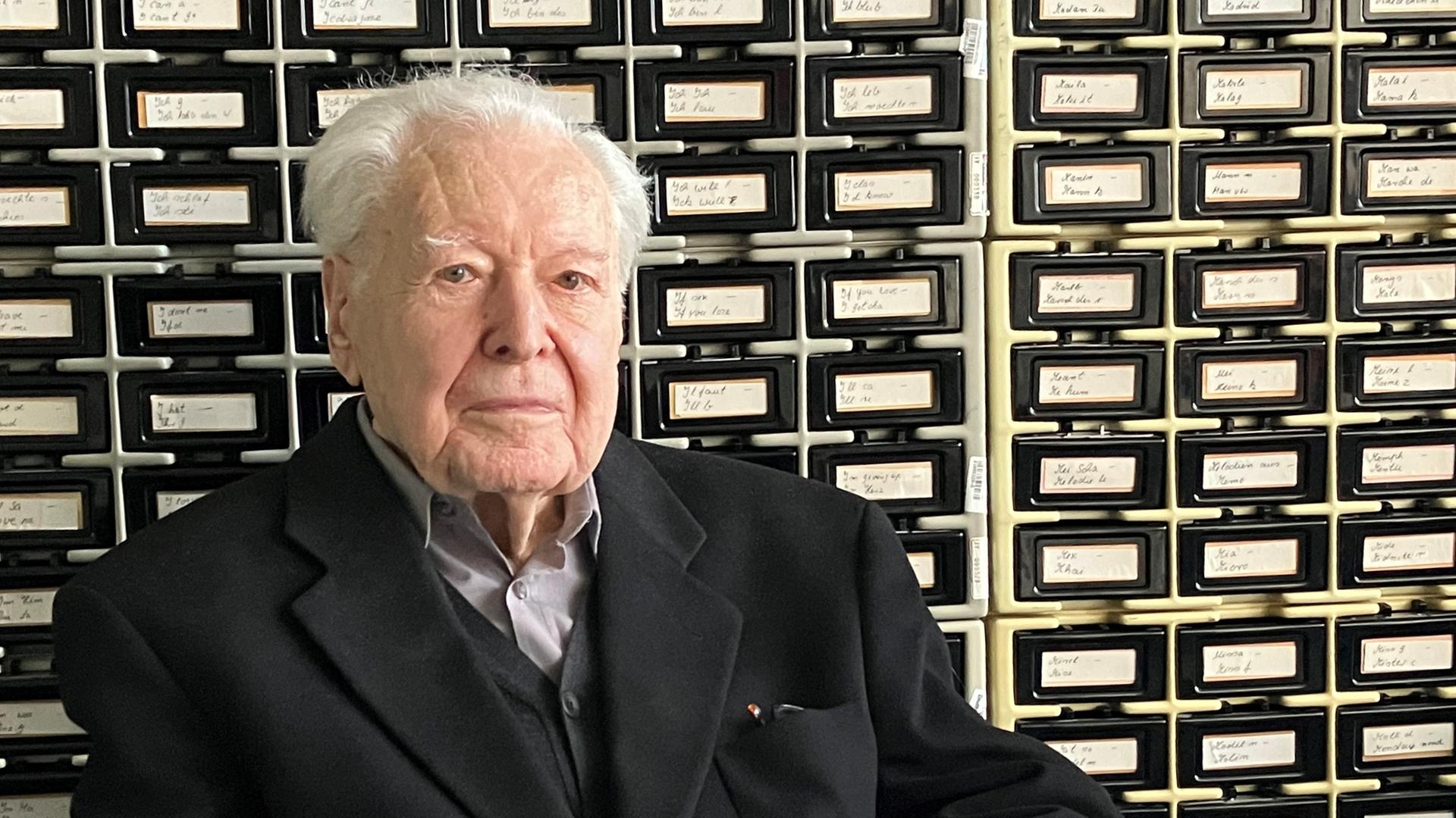 Ein alter Herr mit weißen Haaren sitzt mit gefaltenen Händen vor einem historischen Archivkatalog