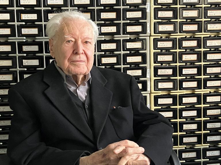 Ein alter Herr mit weißen Haaren sitzt mit gefaltenen Händen vor einem historischen Archivkatalog