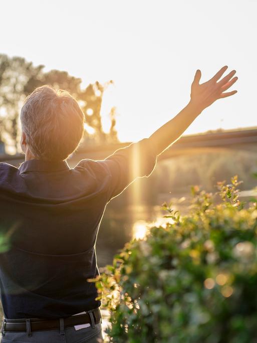 Rückansicht etwas etwas älteren Mannes, der an einem Flussufer steht und mit seinen Armen die Welt zu umarmen scheint. Die Sonne im Gegenlicht verleiht dem Bild eine ekstatische Atmosphäre.