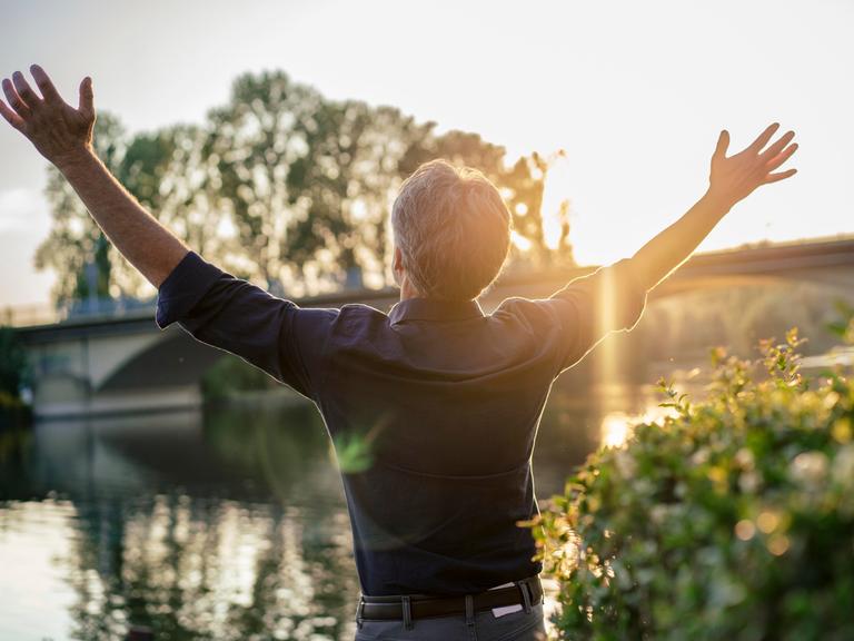 Rückansicht etwas etwas älteren Mannes, der an einem Flussufer steht und mit seinen Armen die Welt zu umarmen scheint. Die Sonne im Gegenlicht verleiht dem Bild eine ekstatische Atmosphäre.