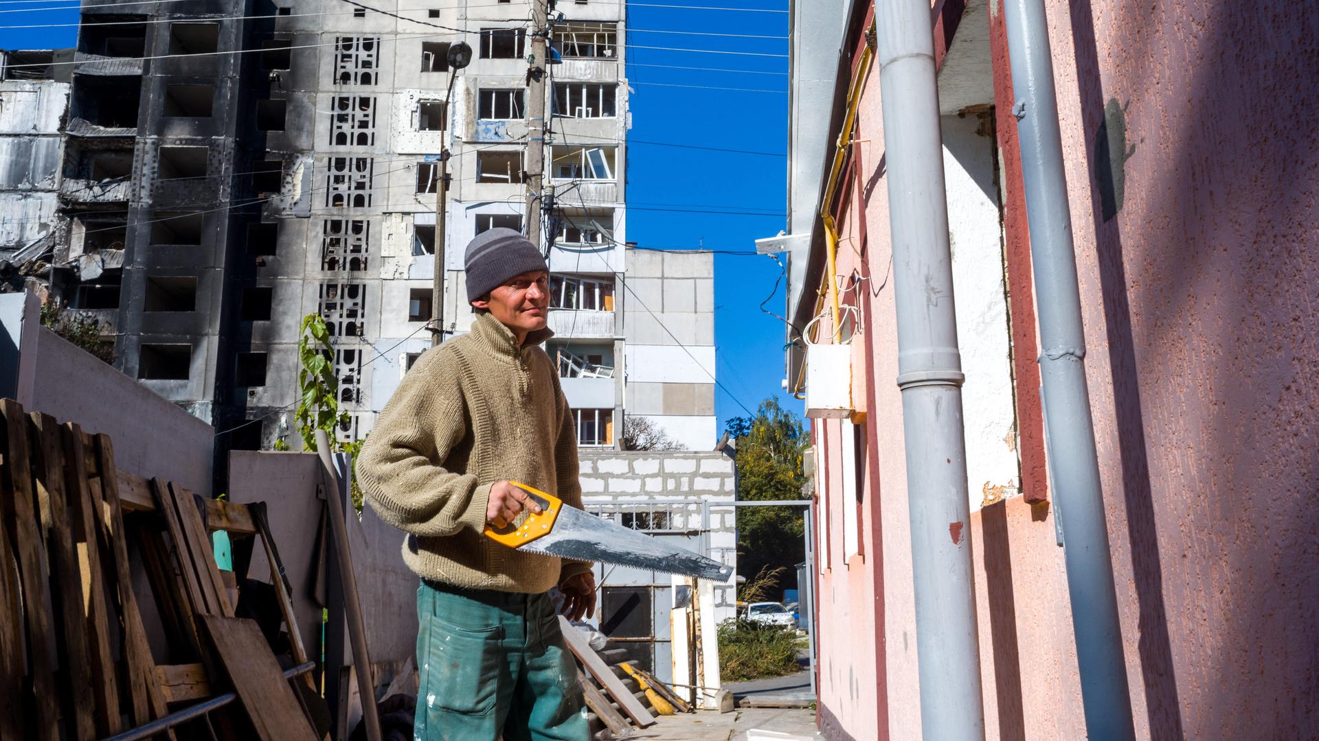 Ein Mann hält eine Säge in der Hand und steht vor einem kriegszerstörten Haus in der Ukraine. 