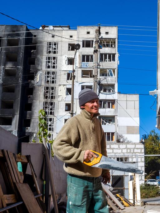 Ein Mann hält eine Säge in der Hand und steht vor einem kriegszerstörten Haus in der Ukraine. 