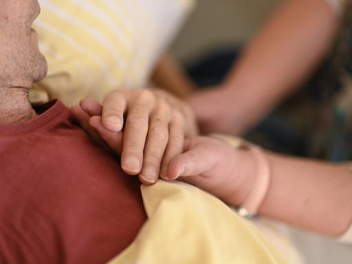 Eine Hospizmitarbeiterin hält die Hand eines todkranken Menschen, der im Hospiz im Bett liegt.