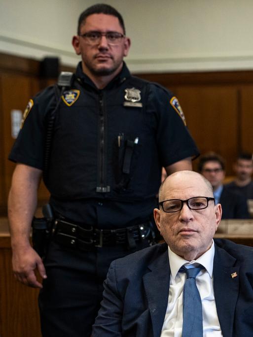 Der Angeklagte US-Filmprozent Harvey Weinstein sitzt, umrahmt von zwei stehenden Polizisten in Uniform.  