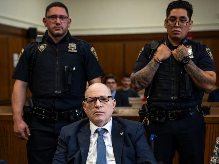 Der Angeklagte US-Filmprozent Harvey Weinstein sitzt, umrahmt von zwei stehenden Polizisten in Uniform.  