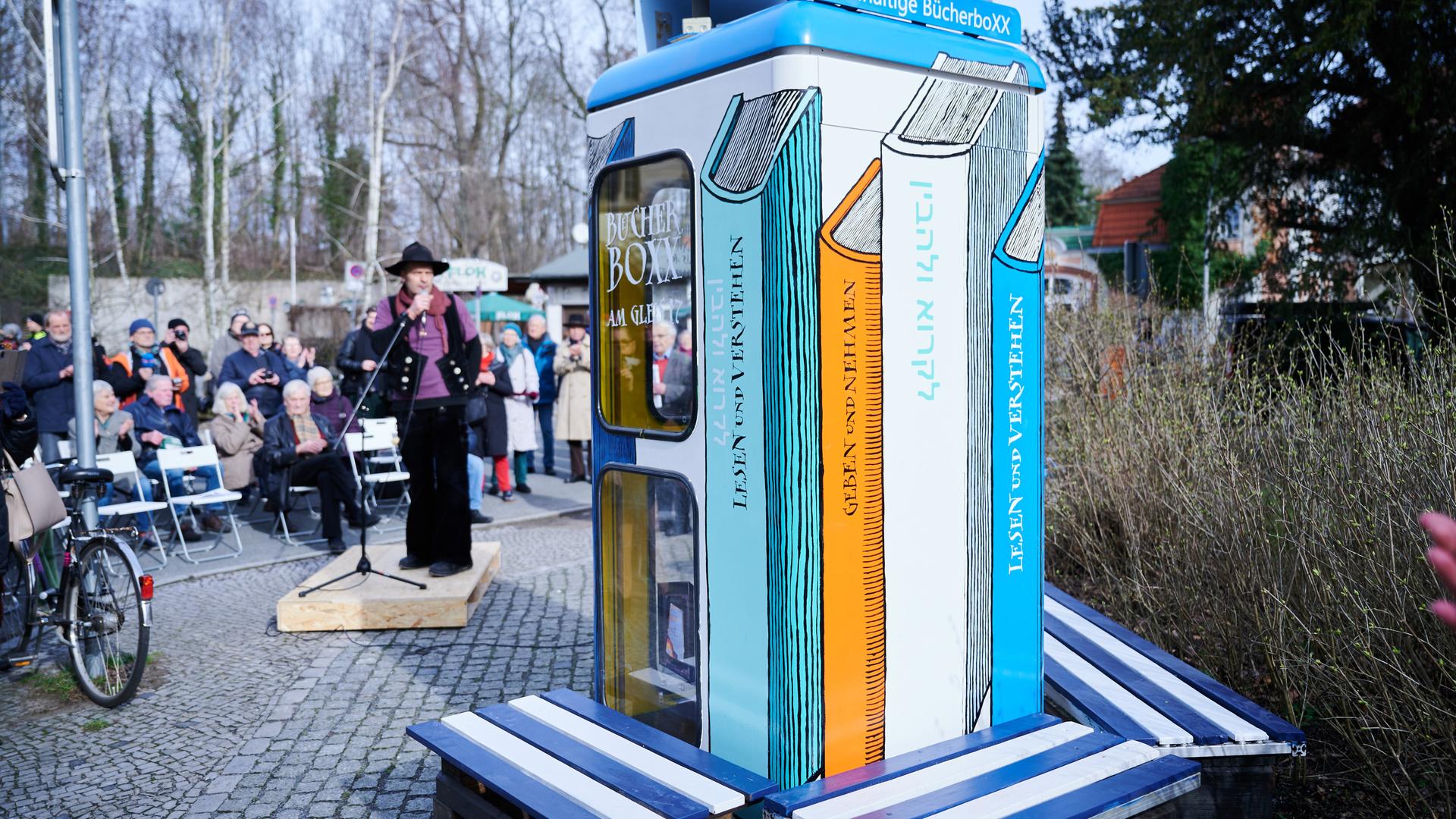 Eine bemalte Telefonzelle steht auf einem Platz. Es ist die Bücherbox am Bahnhof Berlin-Grunewald. Um die Box stehen viele Menschen. Ein Mann hält ein Mikrofon.