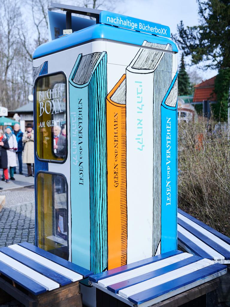 Eine bemalte Telefonzelle steht auf einem Platz. Es ist die Bücherbox am Bahnhof Berlin-Grunewald. Um die Box stehen viele Menschen. Ein Mann hält ein Mikrofon.