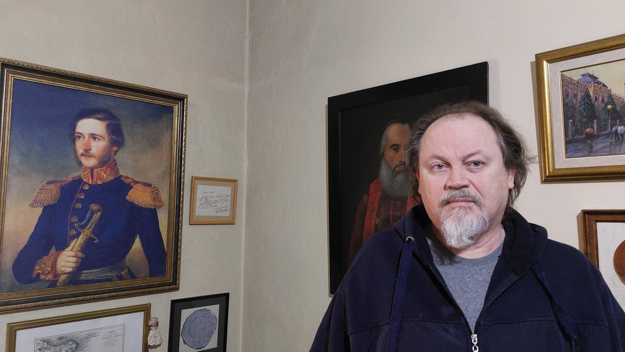 Ein Mann mit weißem Bart steht vor einer Wand mit zahlreichen alten Bildern, einige in Goldrahmen.