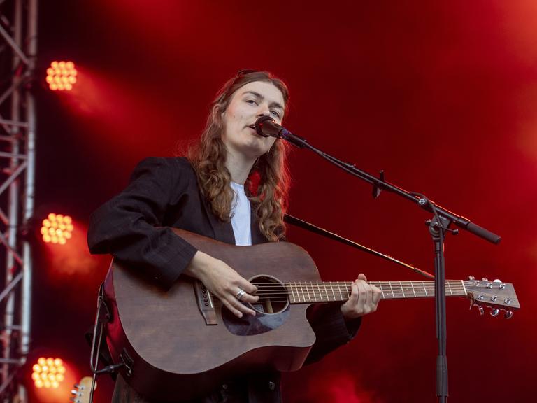 Musikerin Philine Sonny steht bei einem Festival auf der Bühne. Sie singt in ein Mikrofon und spielt dabei ihre Gitarre.