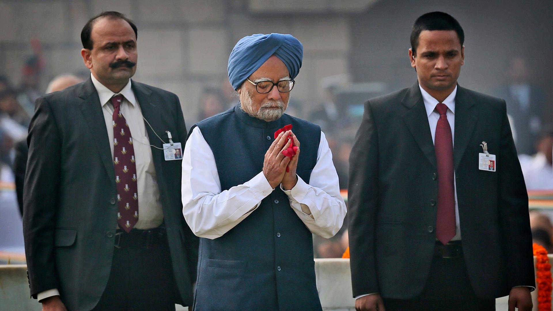 Der frühere indische Premierminister Manmohan Singh steht mit gefalteten Händen vor dem Mahatma-Gandhi-Mahnmal. Neben ihm stehen zwei Männer. 