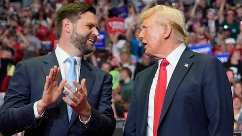 Donald Trump und JD Vance stehen nebeneinander und reden. Im Hintergrund sind viele Menschen zu sehen.
