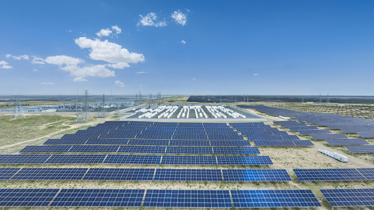 Eine Luftaufnahme zeigt ein weitläufiges Solarkraftwerk mit zahlreichen dunkelblauen Solarmodulen, technischen Anlagen und Strommasten in einer flachen Landschaft unter einem klaren, leuchtend blauen Himmel mit wenigen weißen Wolken.