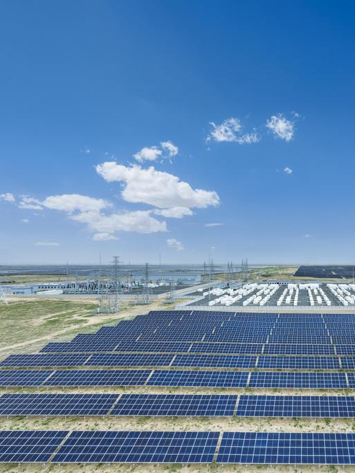 Eine Luftaufnahme zeigt ein weitläufiges Solarkraftwerk mit zahlreichen dunkelblauen Solarmodulen, technischen Anlagen und Strommasten in einer flachen Landschaft unter einem klaren, leuchtend blauen Himmel mit wenigen weißen Wolken. Eine Luftaufnahme zeigt ein weitläufiges Solarkraftwerk mit zahlreichen dunkelblauen Solarmodulen, technischen Anlagen und Strommasten in einer flachen Landschaft unter einem klaren, leuchtend blauen Himmel mit wenigen weißen Wolken.