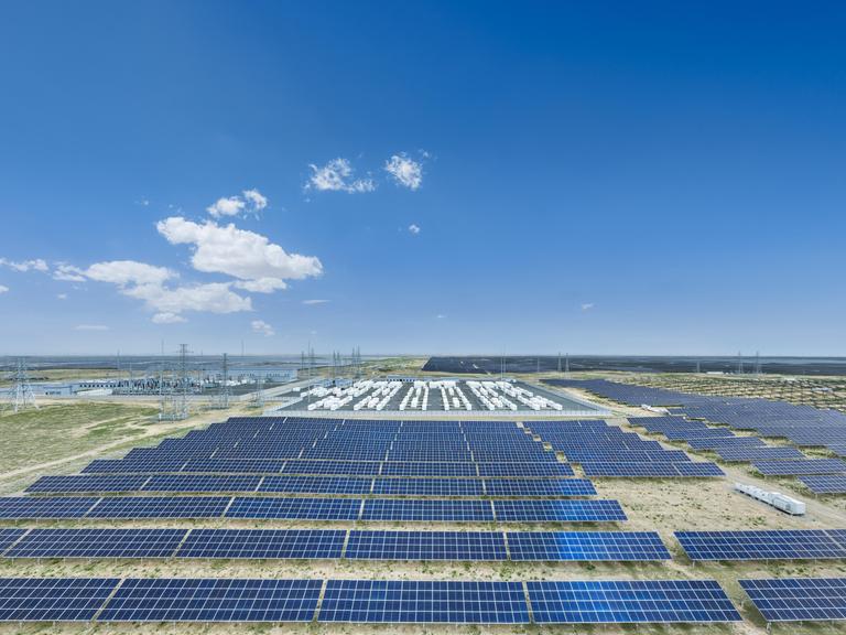 Eine Luftaufnahme zeigt ein weitläufiges Solarkraftwerk mit zahlreichen dunkelblauen Solarmodulen, technischen Anlagen und Strommasten in einer flachen Landschaft unter einem klaren, leuchtend blauen Himmel mit wenigen weißen Wolken. Eine Luftaufnahme zeigt ein weitläufiges Solarkraftwerk mit zahlreichen dunkelblauen Solarmodulen, technischen Anlagen und Strommasten in einer flachen Landschaft unter einem klaren, leuchtend blauen Himmel mit wenigen weißen Wolken.