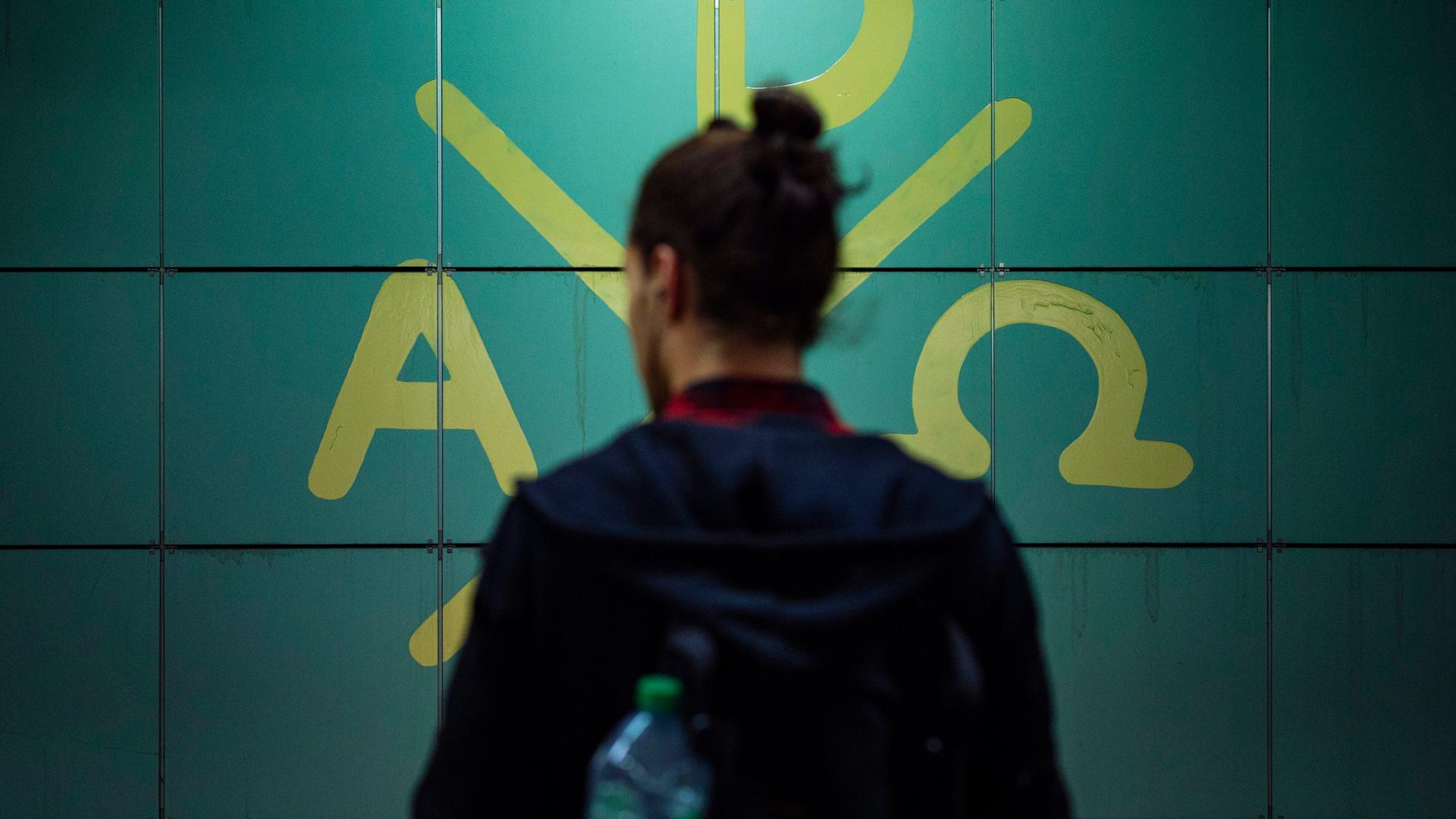 In einem Bahnhof steht ein Fahrgast vor den Symbolen des Christentums. Der Mensch ist nur von hinten angebildet, sein Gesicht ist nicht zu erkennen.