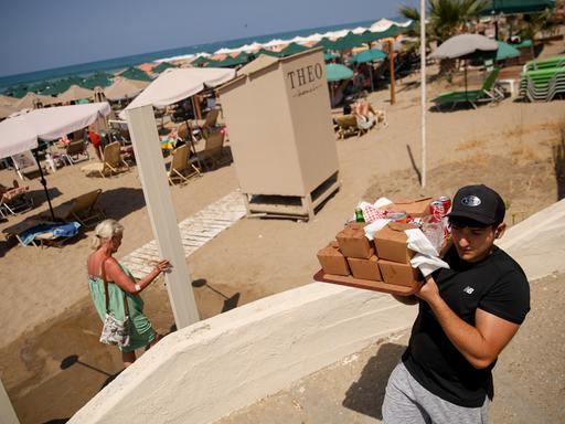 Ein junger Mann in schwarzem T-Shirt und Cap trägt ein Tablett mit Pappschachteln und Getränkedosen auf der Schulter einen Weg entlang. Im Hintergrund ein Strand mit dicht stehenden Sonnenschirmen und Liegen, dahinter das Meer.