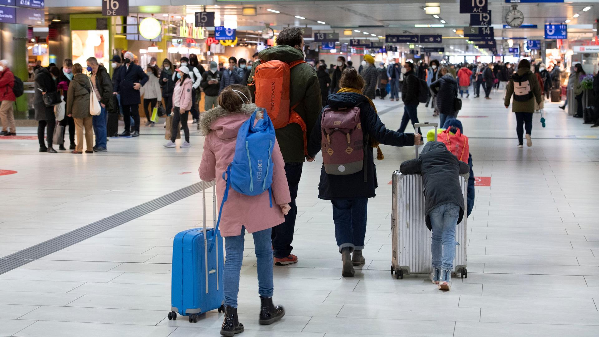 Im Vordergrund einer Bahnhofshalle eine Familie mit zwei Kindern, Rücksäcken und Koffern, im Hintergrund viele Menschen.