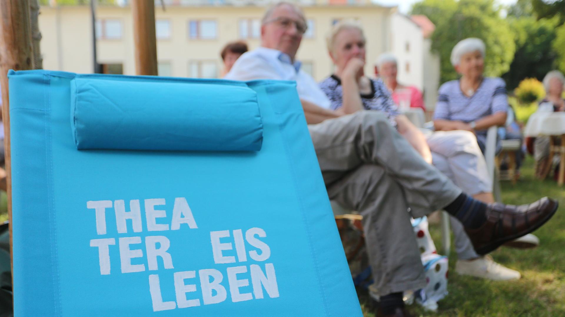 Ein Klappstuhl mit der Aufschrift "Theater Eisleben" steht während eines Picknickkonzertes der Staatskapelle Halle und des Theaters Eisleben auf einer Wiese. Im Bildhintergrund sind Besucher zu sehen.