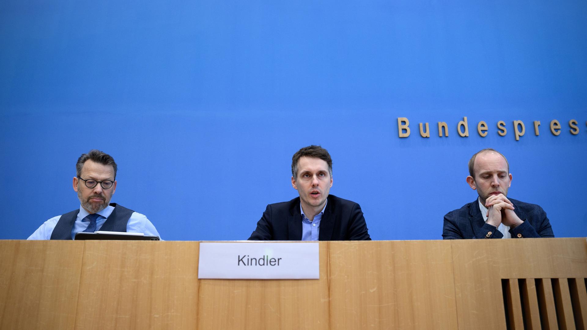 Drei Männer sitzen hinter dem Pult einer Pressekonferenz. Links einer mit Weste und Brille, in der Mitte einer mit Jacket, der gerade spricht und rechts einer mit schütterem Haar, der die Hände wie zum Gebet faltet.