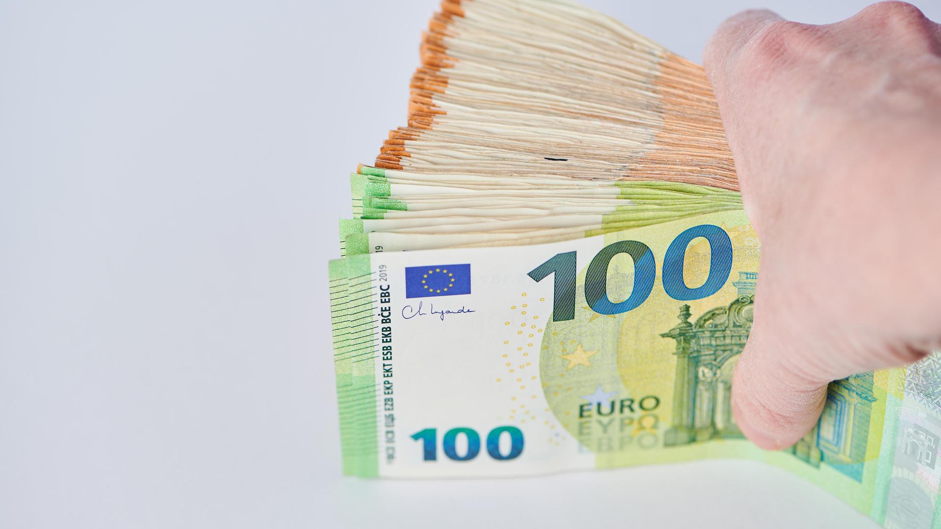 Eine Hand hält eine Bündel Euro-Banknoten vor einem hellen Hintergrund zwischen Daumen und den übrigen Fingern. Eine Hand hält eine Bündel Euro-Banknoten vor einem hellen Hintergrund zwischen Daumen und den übrigen Fingern.