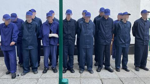 Männer in blauen Arbeitsanzügen und blauen Kappen stehen in zwei Reihen aufgestellt. Ihre Gesichter sind verpixelt.