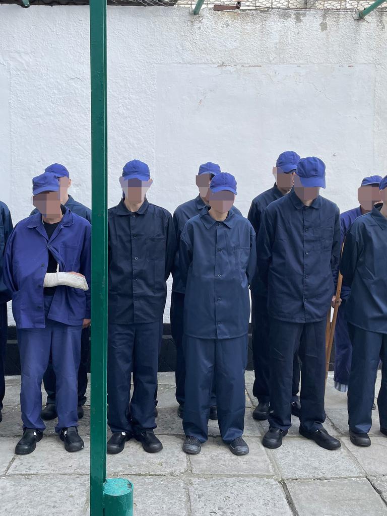 Männer in blauen Arbeitsanzügen und blauen Kappen stehen in zwei Reihen aufgestellt. Ihre Gesichter sind verpixelt.
