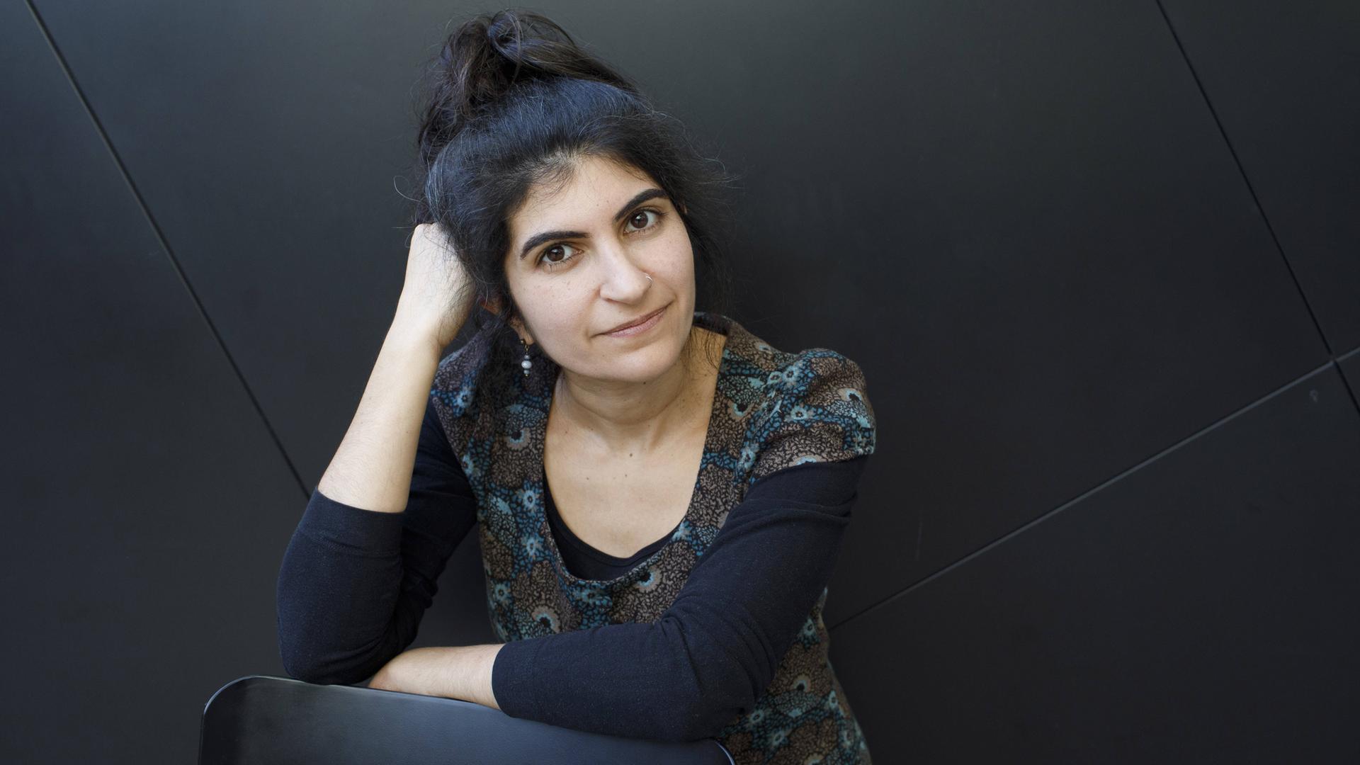 Shida Bazyar sitzt vor einem schwarzen Hintergrund und blickt in die Kamera. Sie hat ihren Kopf auf ihrer rechten Hand aufgestützt