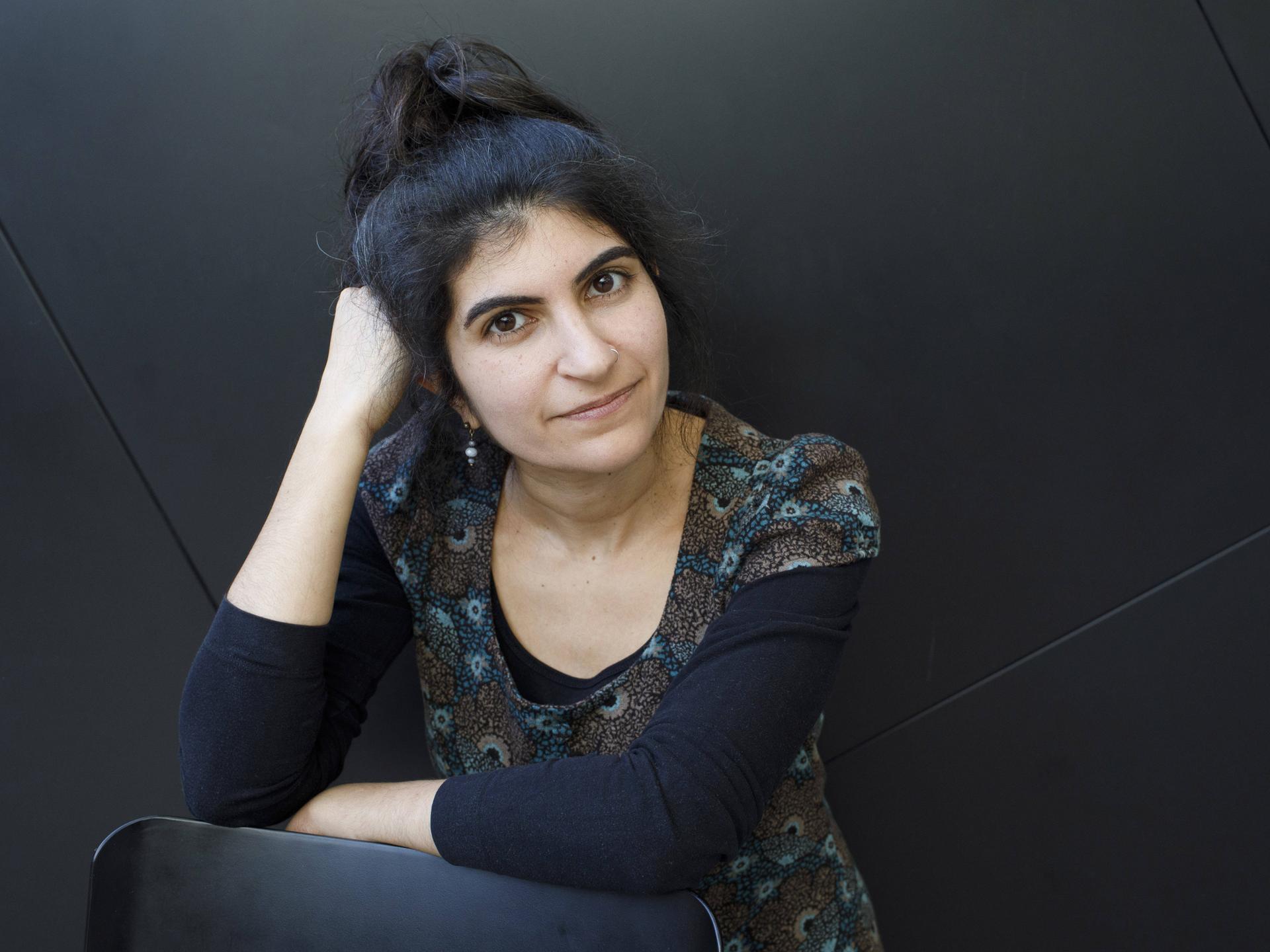 Shida Bazyar sitzt vor einem schwarzen Hintergrund und blickt in die Kamera. Sie hat ihren Kopf auf ihrer rechten Hand aufgestützt