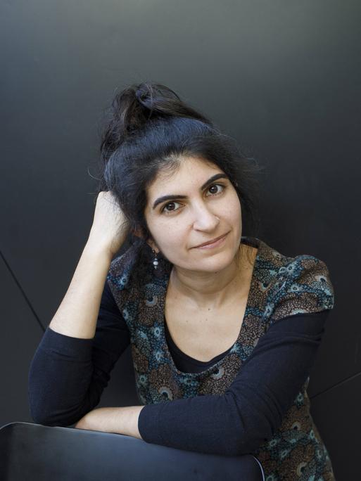 Shida Bazyar sitzt vor einem schwarzen Hintergrund und blickt in die Kamera. Sie hat ihren Kopf auf ihrer rechten Hand aufgestützt Shida Bazyar sitzt vor einem schwarzen Hintergrund und blickt in die Kamera. Sie hat ihren Kopf auf ihrer rechten Hand aufgestützt