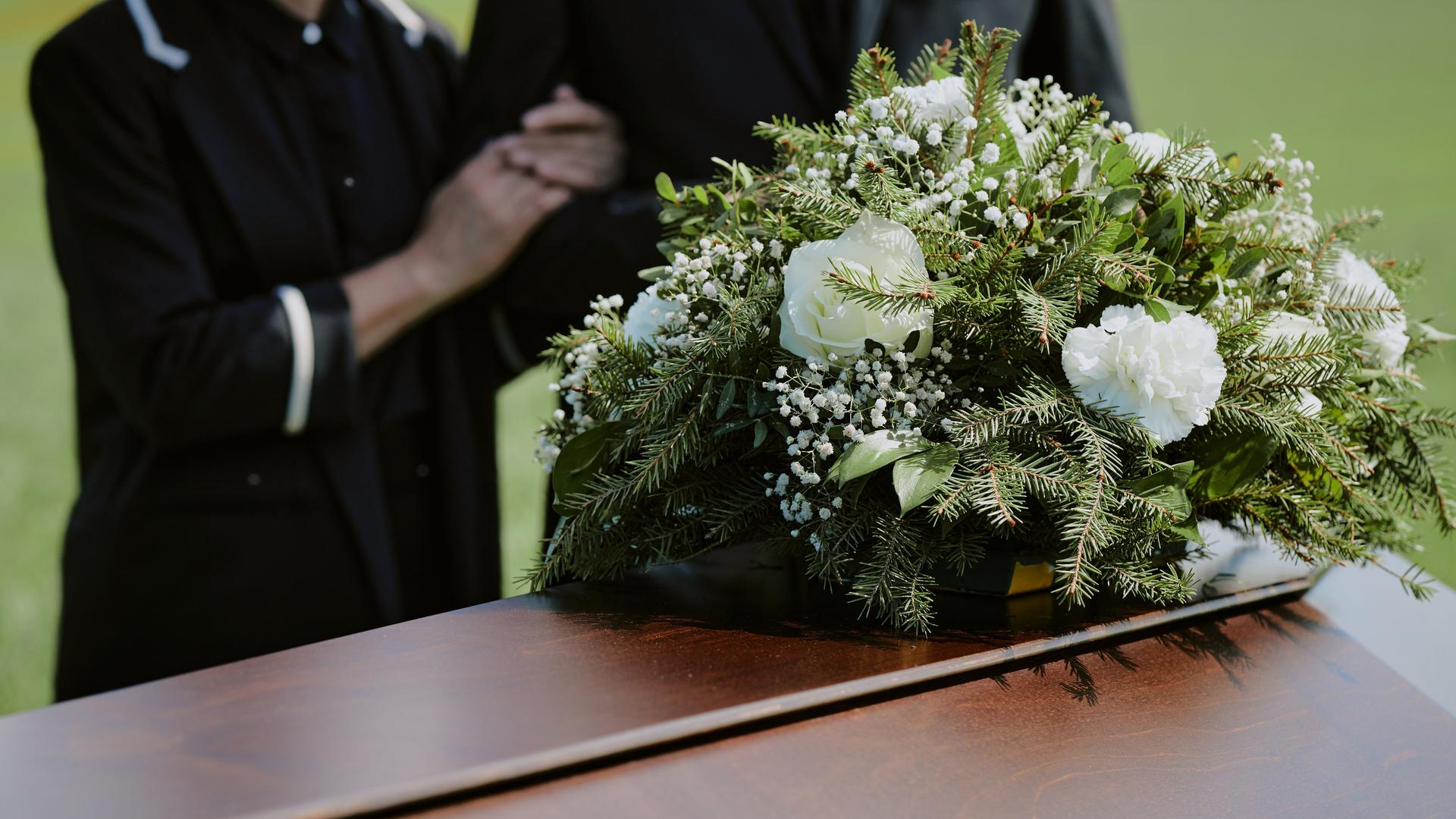 Eine Frau hält den Arm eines Mannes hinter einem Holzsarg mit Blumenschmuck, während einer Beerdigungszeremonie im Freien auf einer grünen Wiese.