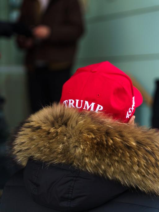 Ein Mann mit einer Jacke mit Fellkragen steht vor einem Haus, er trägt eine rote Mütze mit der Aufschrift "Trump".