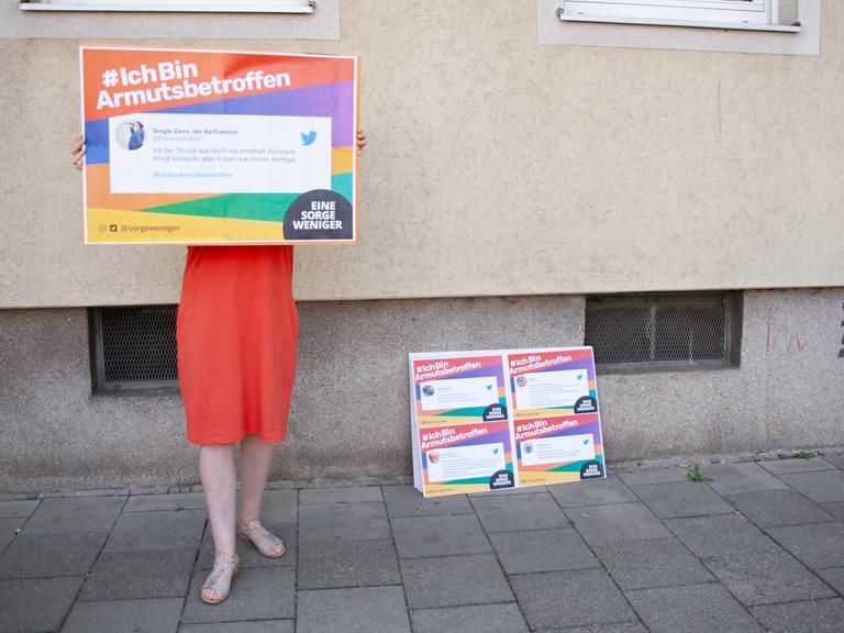 Eine Frau hält ein Schild mit der Aufschrift "#IchBinArmutsbetroffen" und ihrem Tweet dazu vor ihr Gesicht. 