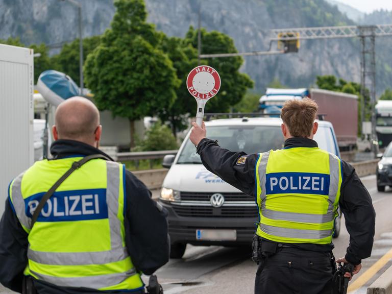Ein Polizist stoppt ein Fahrzeug auf der Autobahn mit einer Polizeikelle mit der Aufschrift: "Halt Polizei". Neben ihm steht ein anderer Polizist.
